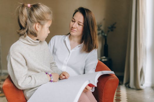 A woman and a child sitting together, reading a Braille book inside a cozy room.