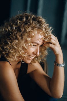 A woman with curly blonde hair and a joyful expression smiles indoors, exuding happiness.