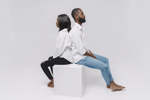 Studio portrait of a couple sitting back to back on a white cube, illustrating relationship dynamics.