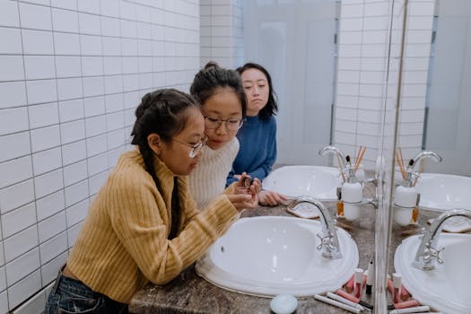 Three young women in a tiled bathroom sharing makeup tips and bonding by the sink.